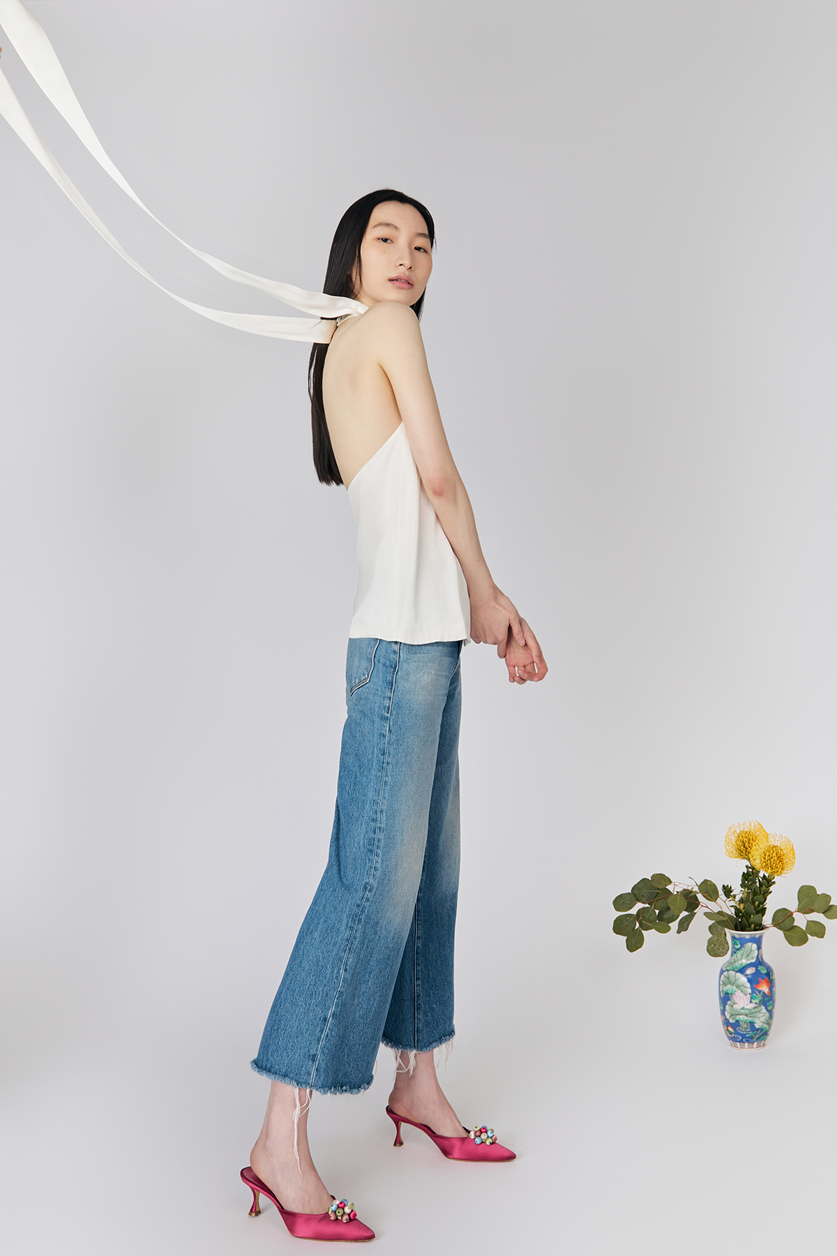 Full side view of model on grey background wearing Du & Berry Moonlight White Silk Halter Top with denim blue jeans showcasing halter tie details flying in the air