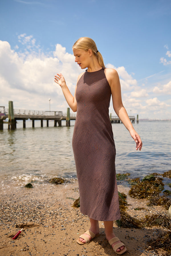 Woman wearing a brown linen side split maxi dress standing on a rocky beach with pier and blue sky