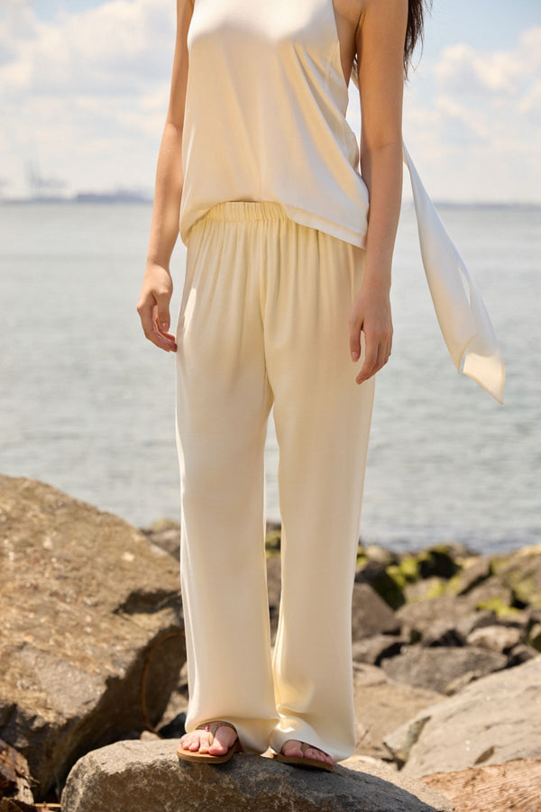 Woman wearing Du & Berry Cream Comfort Straight Pants standing on rocks by the water in a sunny outdoor setting