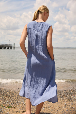 Woman wearing Du & Berry Blue Linen Button Midi Dress standing on a pebbled beach with ocean background