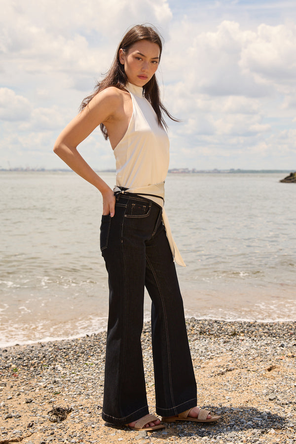Woman wearing navy flare jeans for women from Du & Berry standing on a pebble beach with a sleeveless white top