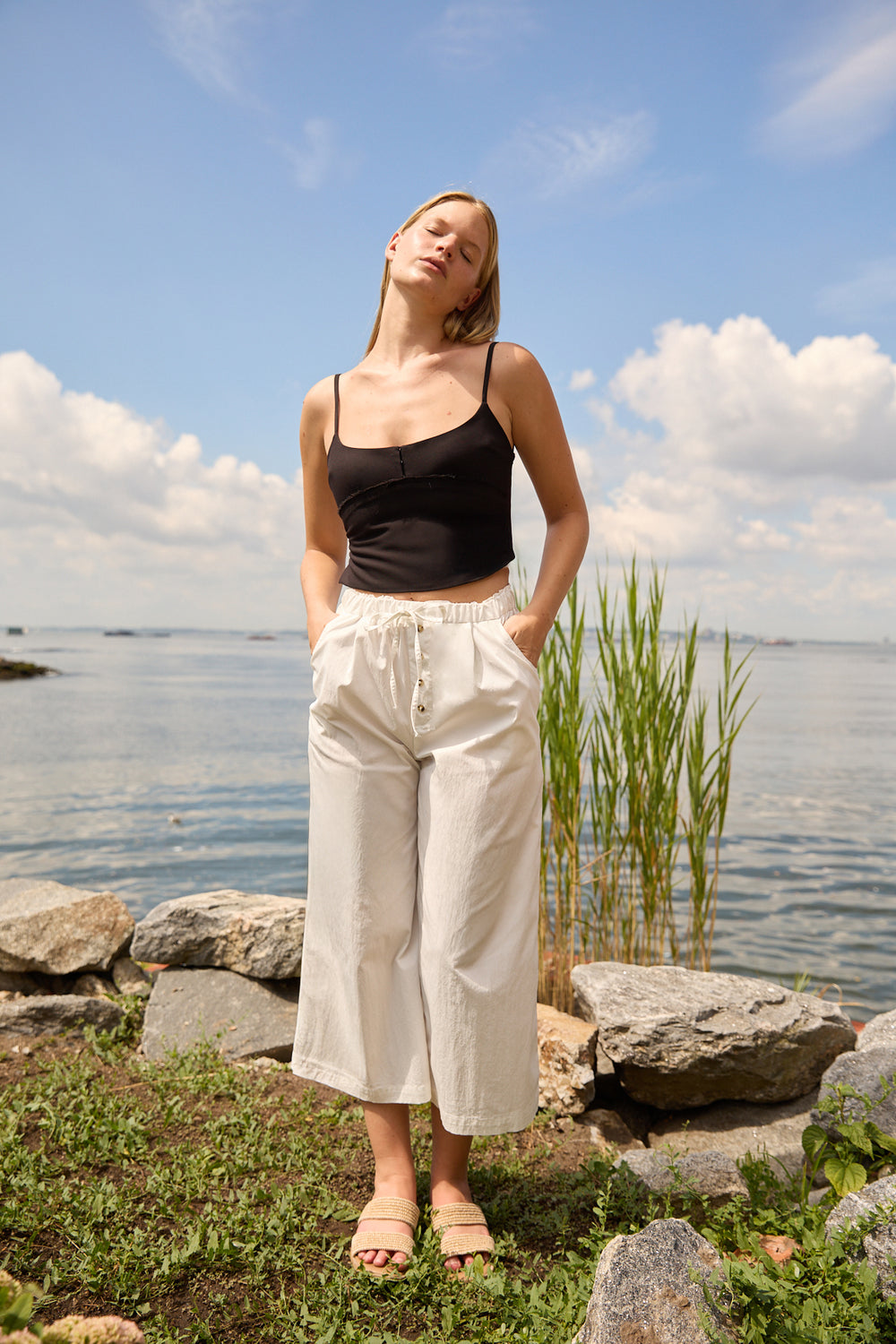 Woman wearing Du & Berry white cotton palazzo pants standing by the rocky lakeside under a blue sky